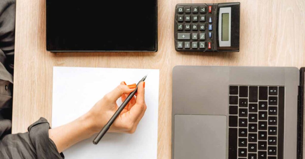 Web developer writing on paper with laptop and calculator around them